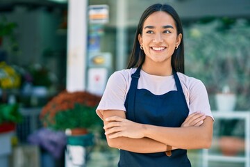 Young latin shopkeeper girl with arms crossed smiling happy standing at the florist - obrazy, fototapety, plakaty