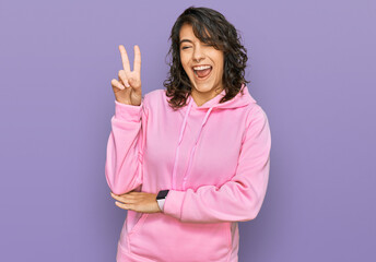 Young hispanic woman wearing casual sweatshirt smiling with happy face winking at the camera doing...