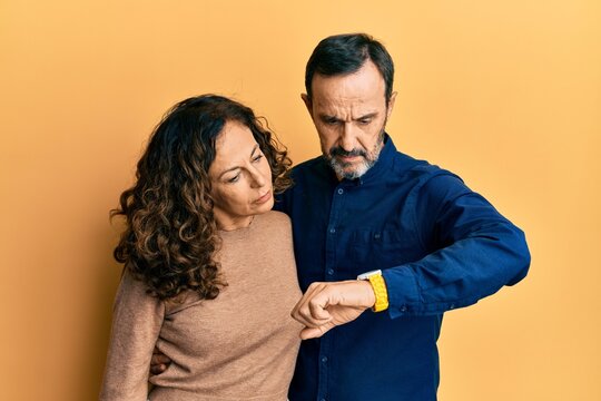 Middle age hispanic couple wearing casual clothes checking the time on wrist watch, relaxed and confident