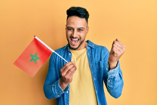 Young arab man holding morocco flag screaming proud, celebrating victory and success very excited with raised arm