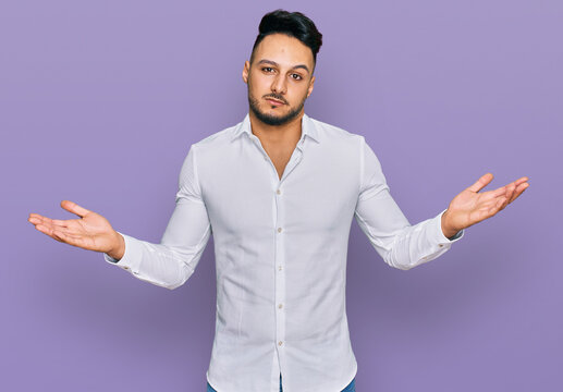 Young arab man wearing casual clothes clueless and confused expression with arms and hands raised. doubt concept.