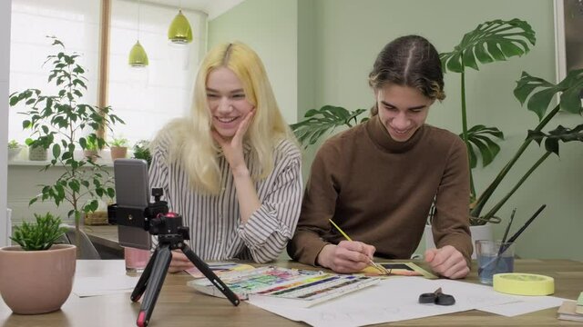 Couple of teenagers painting with watercolors together, guy and girl watching master class on smartphone