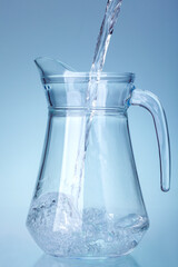 glass transparent jug is filled with a stream of water splashes at the bottom of the container, kitchen utensils isolated on a light cold background.
