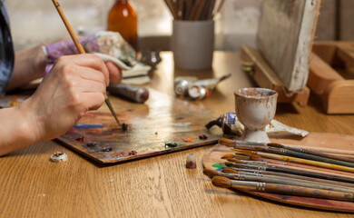 The artist is working on a Still Life. With a brush, he mixes oil paints on the palette.Studio with a desktop easel and brushes on the table. Close-up,Focus on the bunch of brushes in the foreground 