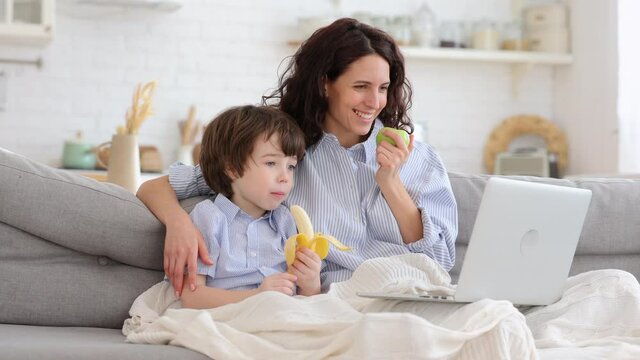 Smiling mother with little son watch funny video, cartoon on laptop computer sit together on sofa in living room. Mom and cute boy with apple and banana fruit under blanket spend time at funny movie