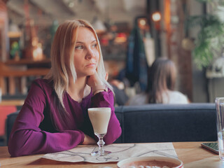 A blonde girl in a lilac sweater sits in a cozy restaurant on a sofa and she is relaxed and smiling and have a coffee drink on her table.