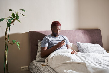 Fototapeta premium Man sitting at the bed after waking up and looking at his smartphone with friendly smile
