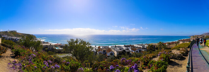 a stunning shot of the vast blue ocean water and a hillside filled with homes and lush green plants and flowers and a footpath with people walking and blue sky at Dana Strands Beach in Dana Point CA - obrazy, fototapety, plakaty