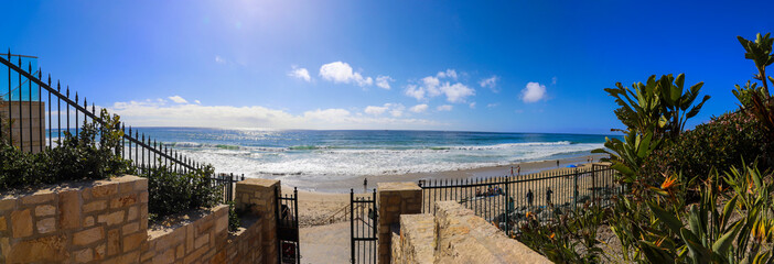 a stunning panoramic shot of the vast blue ocean water with waves rolling in and people walking along the beach on silky brown sand with blue sky and clouds at Dana Strands Beach in Dana Point CA - obrazy, fototapety, plakaty