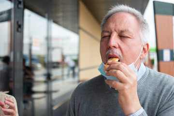 Man eating his fast food lunch hamburger during with a mask on his chin