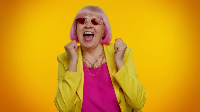 Elderly granny woman in sunglasses and cap listening music, smiling dancing to disco music rhythmically moving her hands, having fun. Senior grandmother isolated on orange background. People emotions