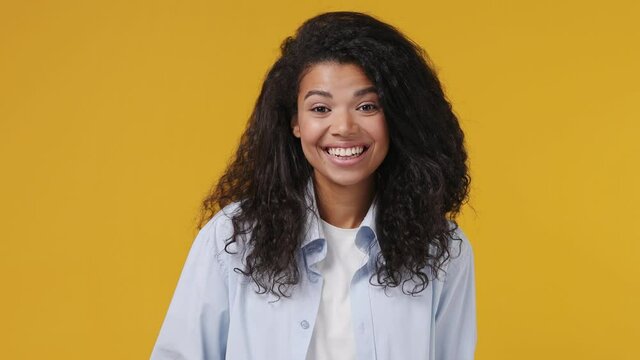 Young fun african woman curly hair 20s years old wears blue white t shirt point finger camera on you doing phone gesture like says call me back oe blink isolated on yellow color wall background studio