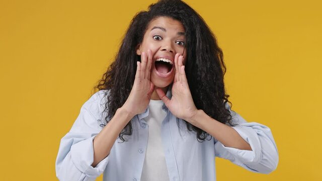 Funny young promoter african woman curly hair 20s years old wears blue white t shirt scream hot news about sales discount with hands near mouth isolated on yellow color wall background studio portrait