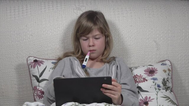 The cute little blonde girl is sick, lies in bed, measures her fever while taking a tablet 