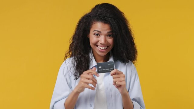 Young african woman curly hair 20s years old wear blue white t shirt pointing index finger on mockup plastic credit bank card showing thumbs up isolated on yellow color wall background studio portrait