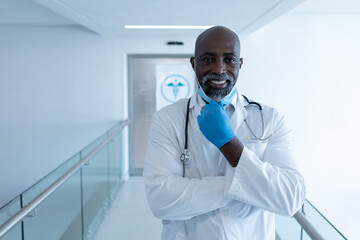 Fototapeta premium Portrait of smiling african american male doctor in hospital corridor holding face mask using tablet