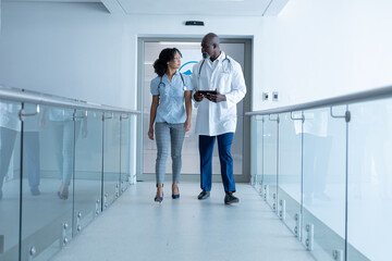 Diverse male and female doctor walking in hospital corridor talking and looking at tablet