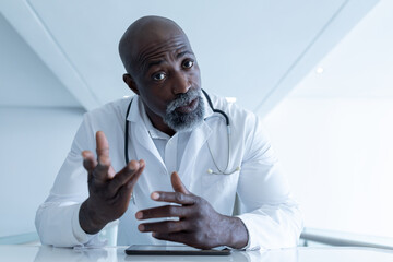 African american male doctor sitting at desk talking and gesturing during video call consultation