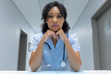 Mixed race female doctor at desk leaning chin on hands and listening during video call consultation