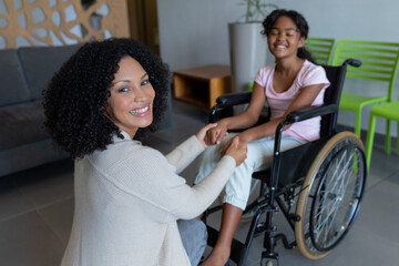 Happy mixed race mother kneeling holding hands with smiling daughter in wheelchair in hospital foyer