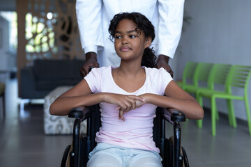 African american male doctor pushing smiling mixed race girl in wheelchair through hospital corridor
