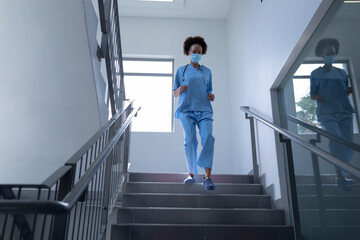 Mixed race female doctor wearing face mask and scrubs running down stairs in hospital