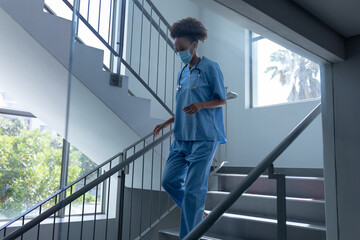 Mixed race female doctor wearing face mask and scrubs walking down stairs in hospital