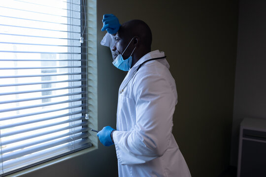 Thoughtful African American Male Doctor In Hospital With Face Mask Looking Out Of Window