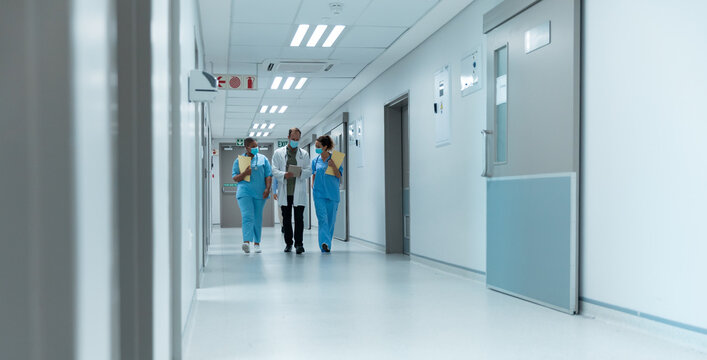 Diverse Group Of Male And Female Doctors Wearing Face Masks Walking Looking At Tablet