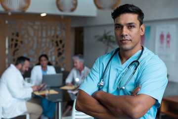 Portrait of mixed race male doctor with arms crossed, colleagues in discussion in the background