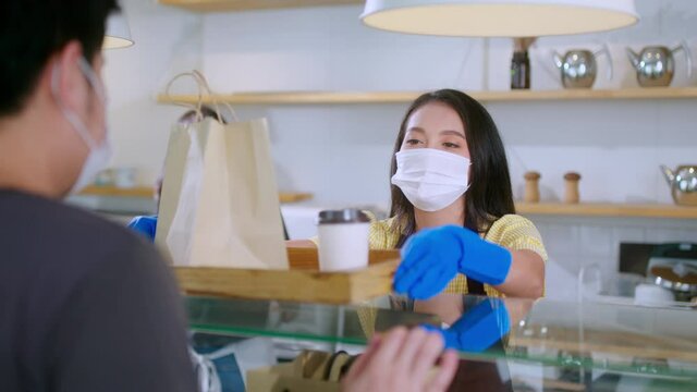 Attractive asian female cafe worker wears face mask and gloves giving takeaway food bag to customer.waitress holding takeout order standing in coffee shop front door entrance new normal lifestyle