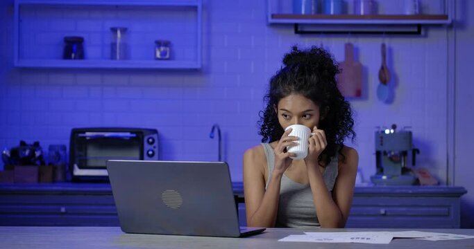 A young, exhausted and tried Asian woman sitting in kitchen and drinking coffee while making video call with her co-worker to finishing important project. Working late at night and New normal concept.