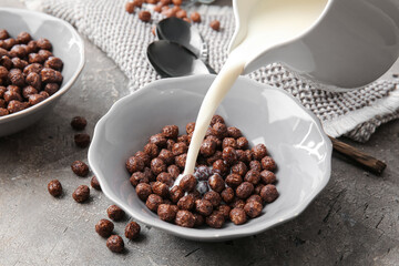 Pouring of milk into bowl with chocolate corn balls on grey background