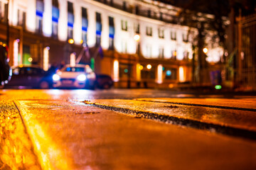 Obraz premium Autumn rainy night in the city. Headlights of a riding car. Parked cars. Residential buildings in the city center. Colorful colors. Close up view from the level of a pedestrian crossing.