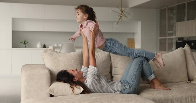 Mother lifts on arms her cute preschool daughter, kid spread arms on the sides imagining flying on air pretend to be airplane have fun play with loving mom while she lying on comfy sofa in living room