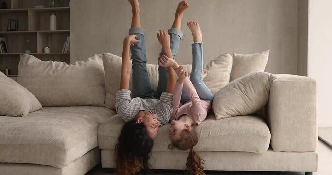 Joyful mom little daughter fooling while lying upside down on sofa. Playful family cheerful woman and preschool girl have fun jerk with raised arms and legs. Funny weekend leisure with child concept