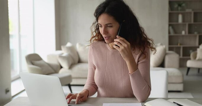 Woman sit at desk in modern living room talk on phone receive or provide consultation remotely, make order distantly. Businesswoman looks at laptop screen chat with colleague discuss business concept
