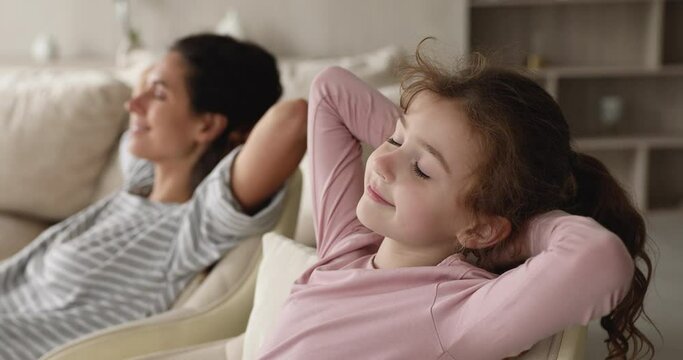 Calm little daughter relax with mom leaned on cozy sofa, close up focus on kid. Peaceful family closing eyes put hands behind head enjoy fresh conditioned air inside of modern flat. No stress concept
