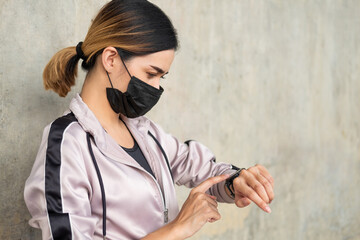Selective focus at face of young beautiful Asian women wearing surgical face mask using smart watch to track activity before exercise or running at the park in the morning. New normal lifestyle.