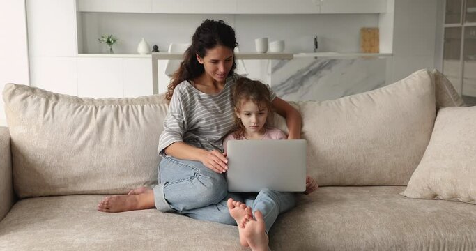 Young mom little daughter sit on sofa in living room with laptop use modern tech for studying child development or fun, choose goods on e-commerce retail services, spend weekend using internet concept