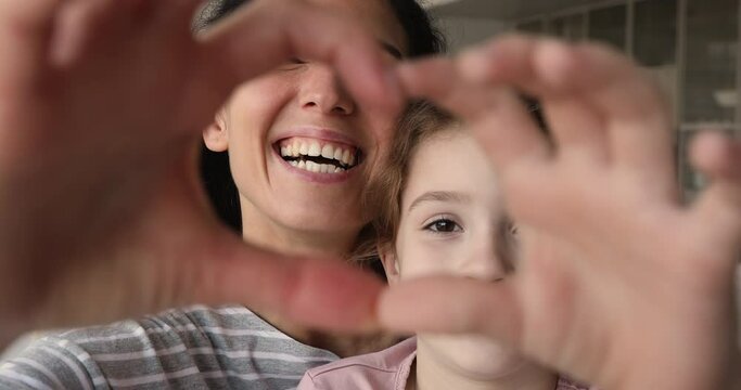 Beautiful happy mother and little cute daughter faces seen through joined fingers making heart shape, close up. Symbol of love, cherish, family bonding, donation sign, adoption and custody concept