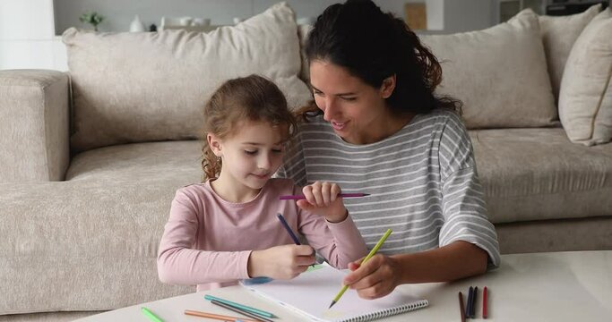 Young mother and little cute preschool daughter sit at table in cozy living room chatting enjoy drawing with pencils in sketchbook, family hobby on weekend at home, child development, pastime concept