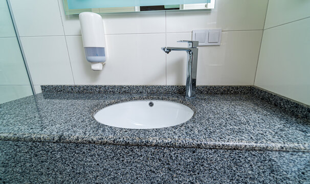 Public Clinic Bathroom With Soap Dispenser On Tiled Wall With Mirror. Granite Sink For Washing Hands And Face. Closeup.