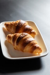 French croissant on white plate on black table background and nature sunlight with shadow through from window.
