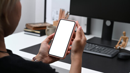 Close up over shoulder view of young woman sitting in front of computer and using mobile phone.
