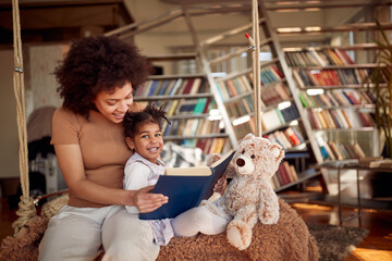 A young mother loves reading a book to her little daughter while sitting on the swing at home together. Family, together, leisure