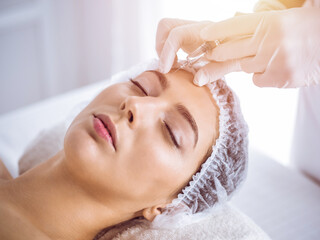 Obraz na płótnie Canvas Beautician doing beauty procedure with syringe to face of young brunette woman in sunny spa center. Cosmetic medicine and surgery, beauty injections