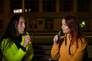 Two caucasian women smoke vape and hookah outdoors at night.