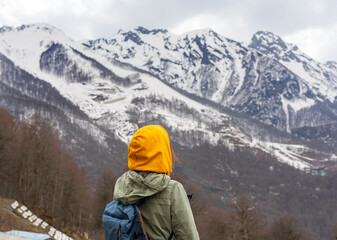 Fototapeta na wymiar Young woman in yellow hood with backpack from behind enjoying the view of the Caucasian mountain peaks covered with snow, hiking, active lifestyle, landscape