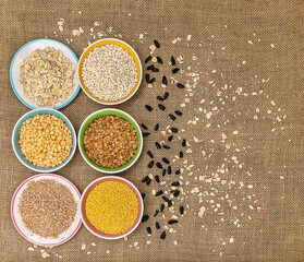 Portions of various cereals - wheat, peas, buckwheat, millet, oatmeal, barley in a multi-colored dish against a background of burlap and scattered sunflower seeds and flakes, flat lay, copy space